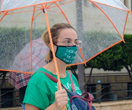 A woman in Almería wearing a face mask stands under an umbrella during rainfall.