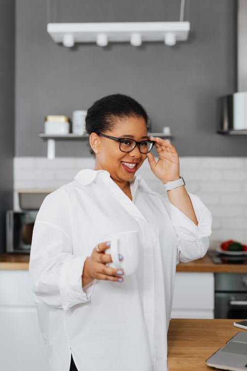 Free Happy woman in a white shirt holding a mug, standing in a modern kitchen. Stock Photo