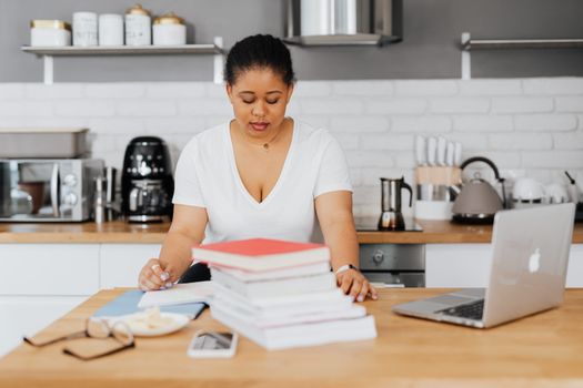 Adult woman focusing on studying in a contemporary kitchen space with books and laptop.