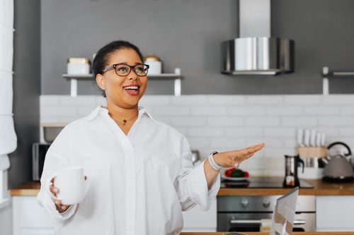 Free Cheerful African American woman in a kitchen with a mug and laptop, showing positive emotion. Stock Photo