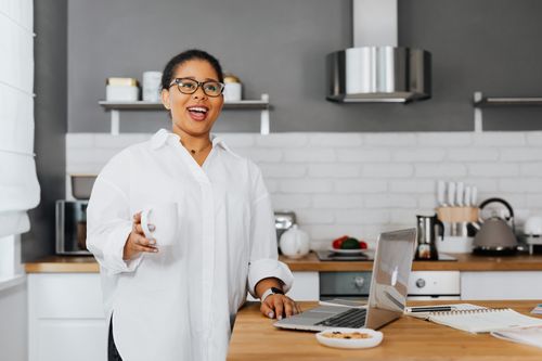 Free Confident woman in glasses holding a mug, smiling while standing in a modern kitchen with a laptop. Stock Photo