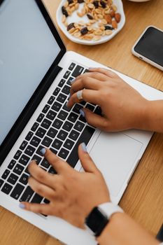 Close-up of hands typing on a laptop with a smartphone and a saucer of mixed nuts on a wooden table.