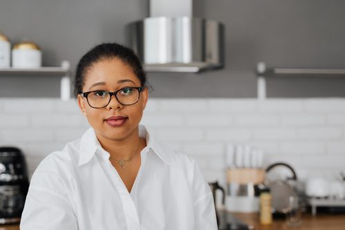 Free Portrait of a black woman in a modern kitchen, wearing eyeglasses and a white button-up shirt. Stock Photo