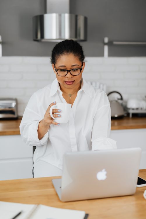 Free Professional woman in white shirt working on laptop while enjoying a drink in a stylish kitchen. Stock Photo