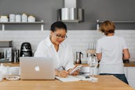 一位成年女性在时尚的厨房环境中，一边写作一边使用笔记本电脑，在现代氛围中处理多项任务