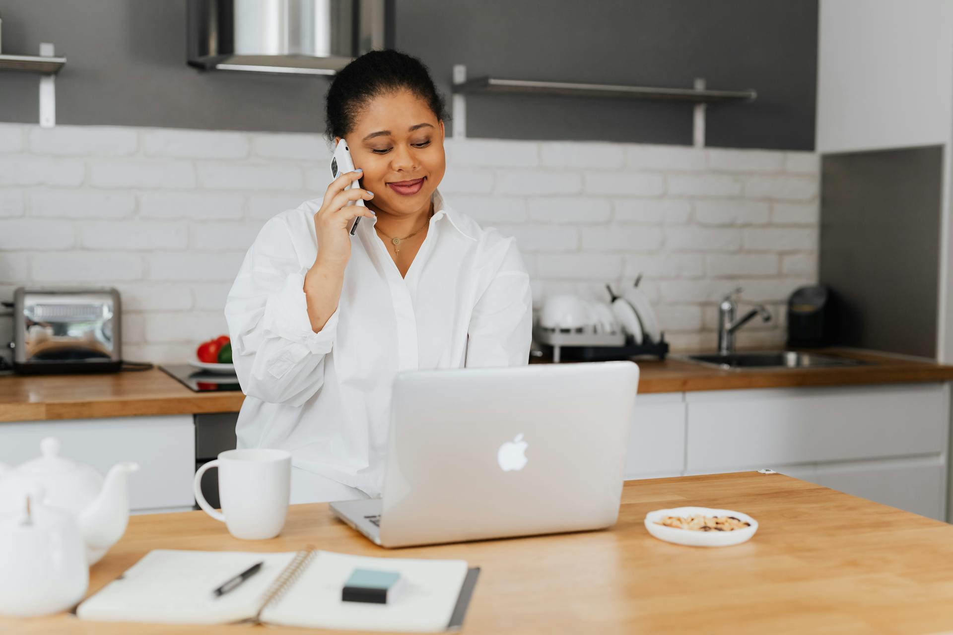 African American woman in white shirt at home desk using phone and laptop.