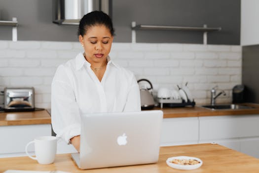 A woman focuses on working on her laptop in a stylish kitchen setting.
