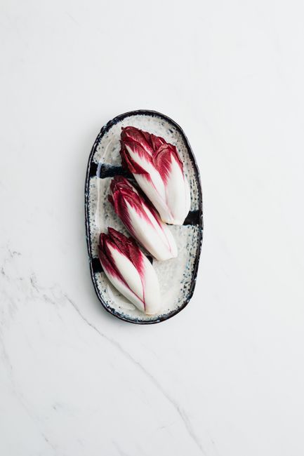 Three pale yellow-white Belgian endive heads with tightly furled leaves resting on a wooden cutting board next to a halved endive showing its compact core.
