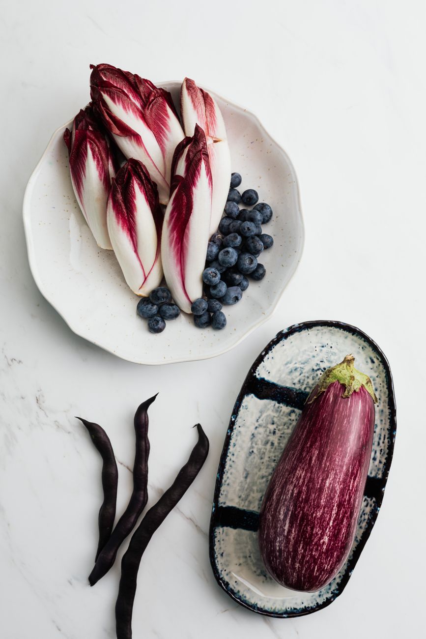 Colorful display of endives, eggplant, blueberries, and beans on a marble surface.
