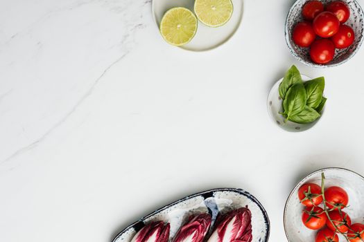 Top view of fresh tomatoes, limes, basil, and radicchio on a marble surface.