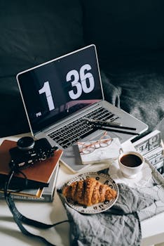 A cozy home office setup with a laptop, coffee, croissant, and books on a sofa.