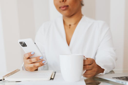 A woman wearing a white shirt using a smartphone while holding a coffee mug indoors.