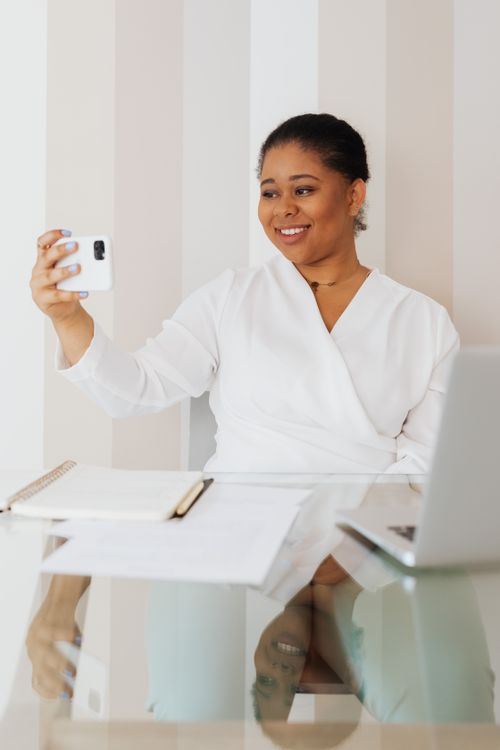 Free Smiling woman in white blouse taking a selfie while sitting at a glass desk in an office setting. Stock Photo