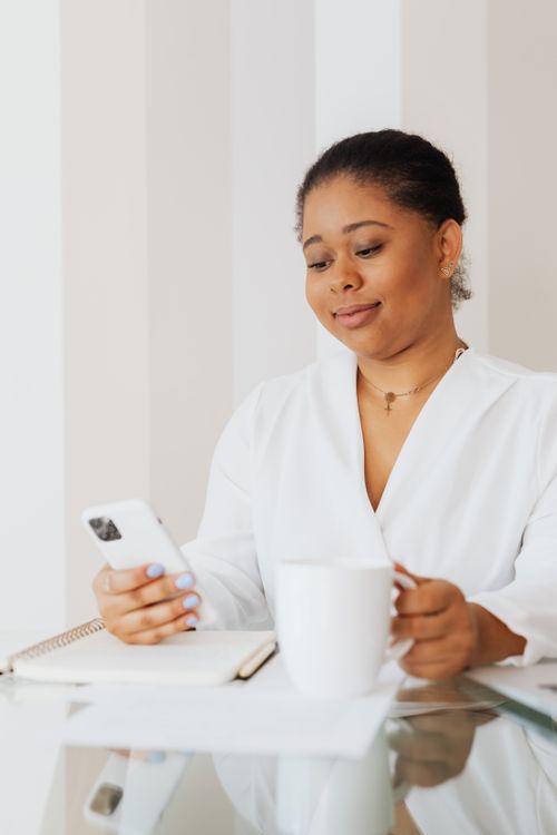 Free African American woman using smartphone with a coffee mug indoors, showcasing modern technology and relaxation. Stock Photo