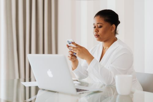 Free A woman in white attire uses her smartphone while working on a laptop at a desk indoors. Stock Photo