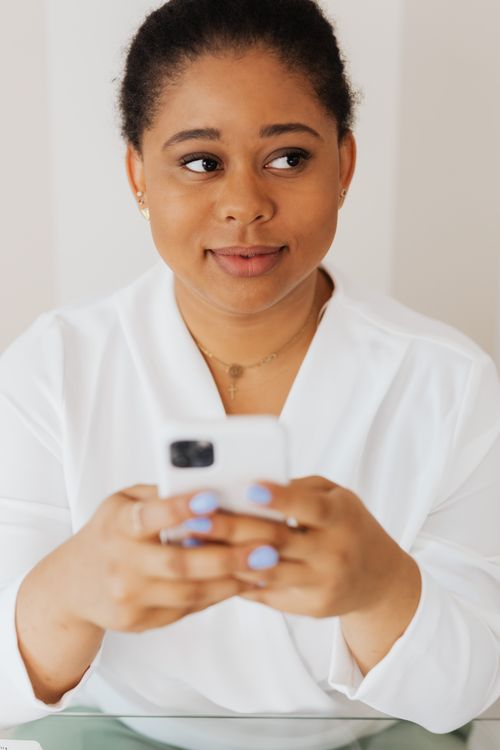 Free Black woman holding smartphone, wearing white blouse, in a contemplative mood. Stock Photo