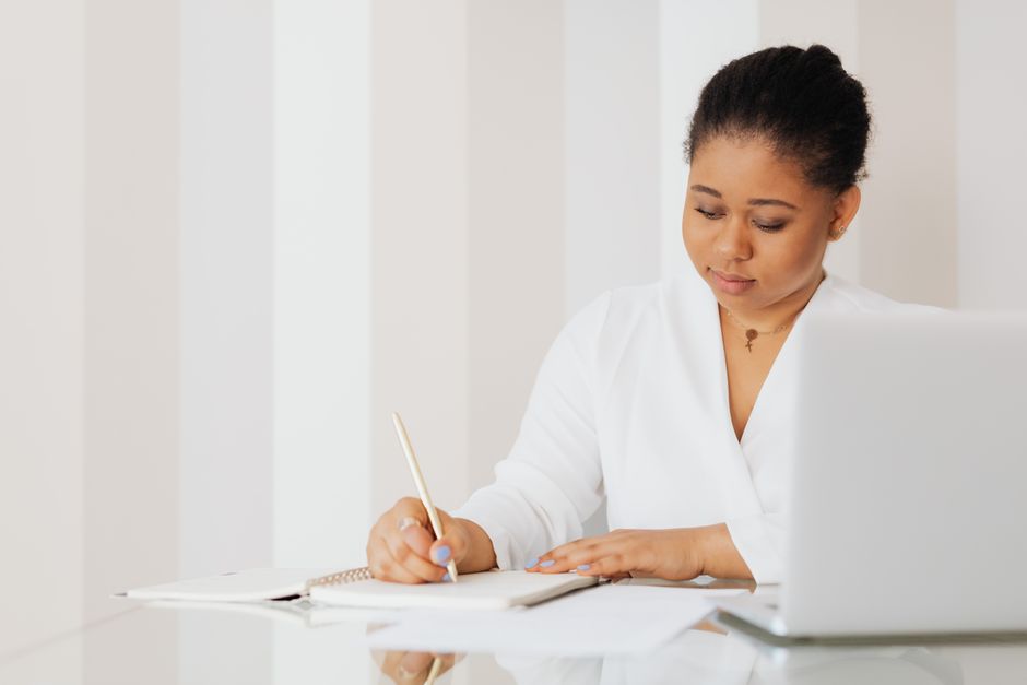 Woman in white top writing notes at a desk with laptop. Ideal for business and productivity themes.