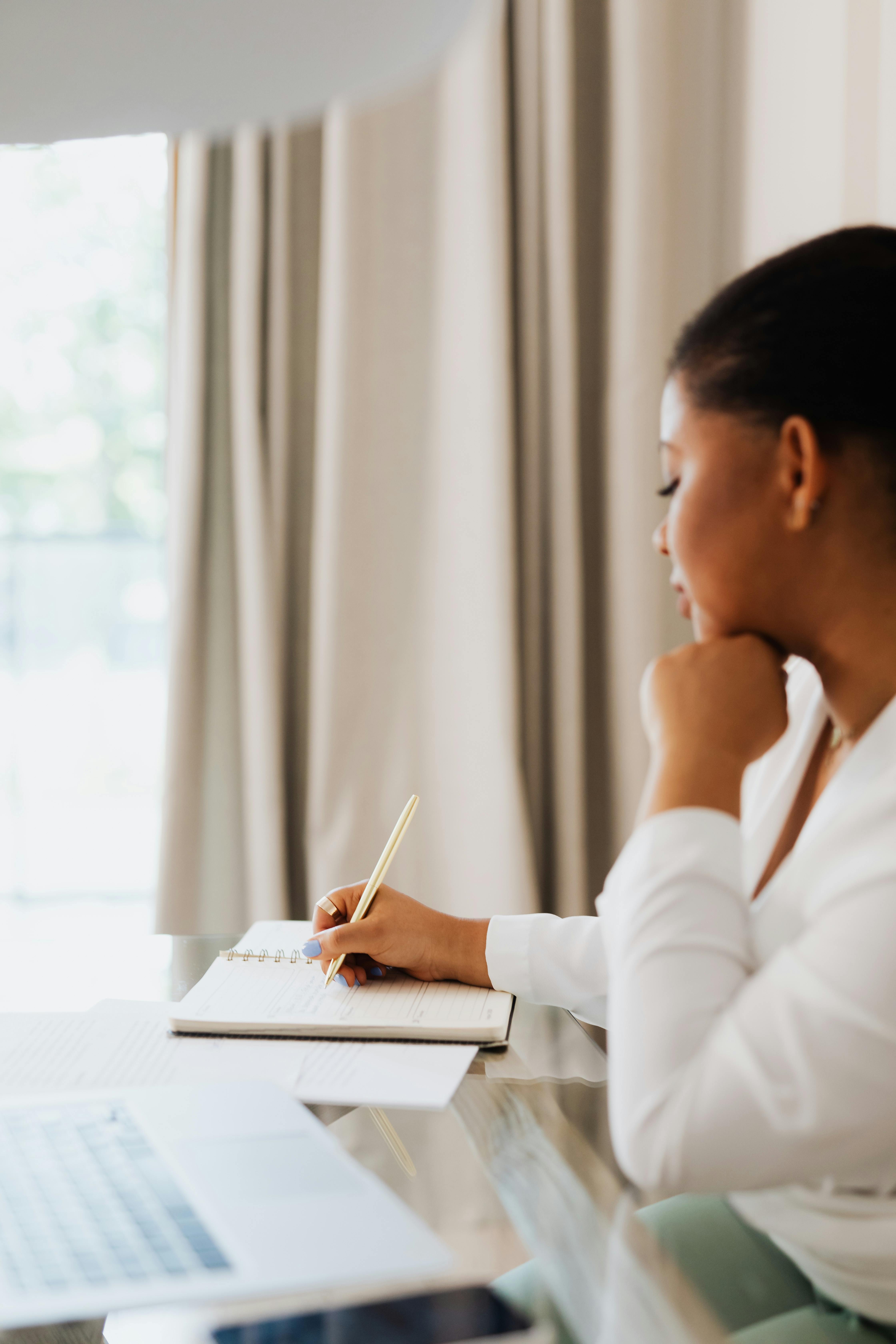 Free A woman in a white shirt sits indoors by a window, writing notes in a notebook. Stock Photo