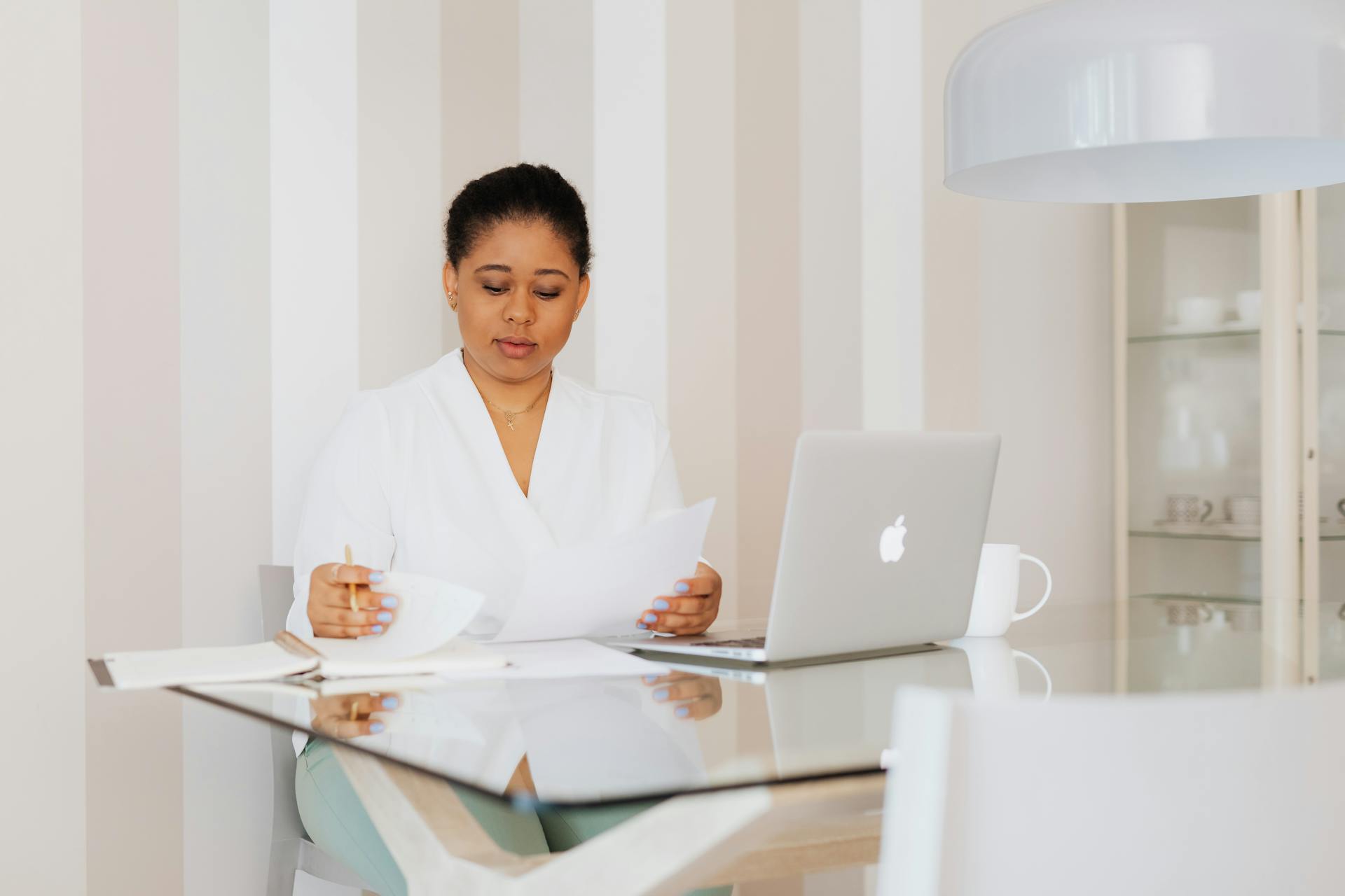 Businesswoman in a modern office, reviewing documents at a glass desk with a laptop.