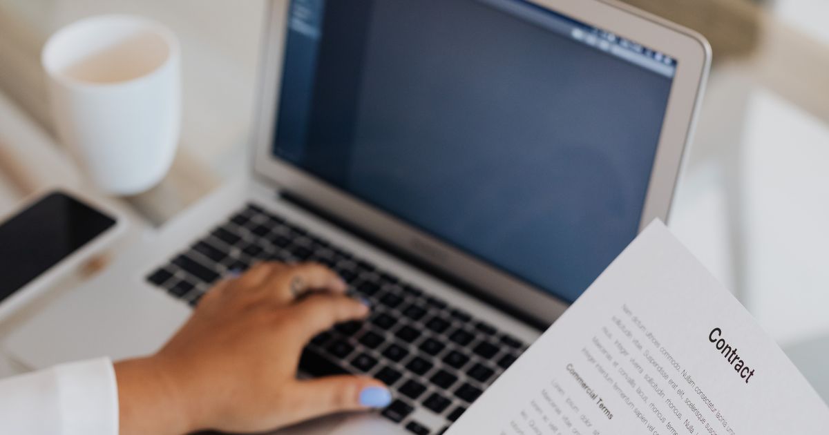Une main tient un contrat imprimé devant un ordinateur portable ouvert, l'autre main posée sur le clavier, une tasse et un téléphone sur le bureau.