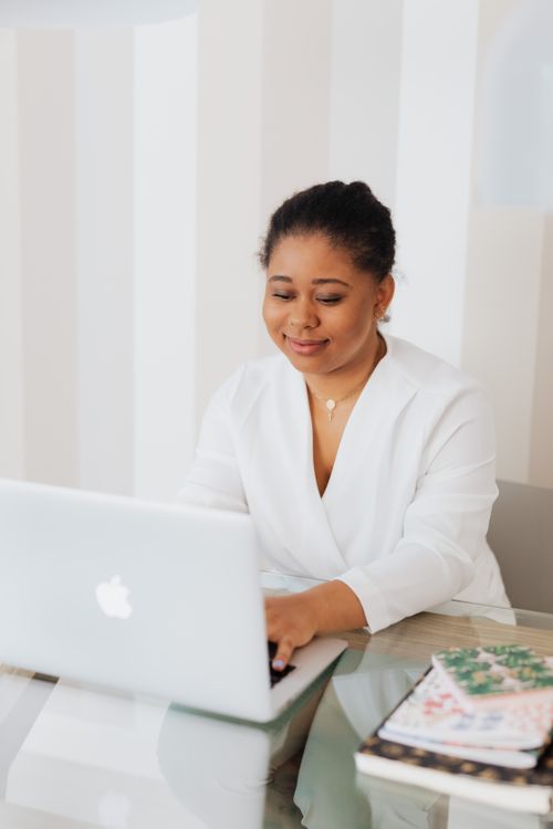 Free Smiling black woman in an office setting, wearing white, using a laptop at a tidy desk. Stock Photo
