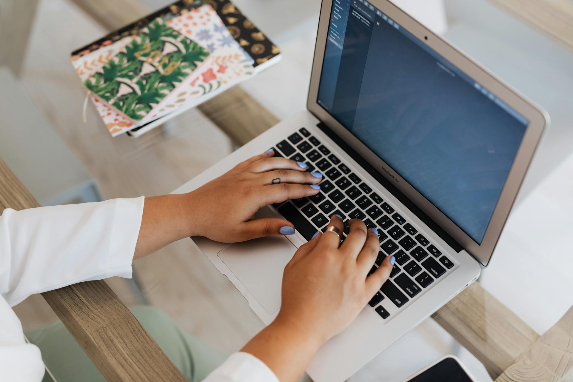 Close-up of a woman's hands typing on a laptop with notebooks nearby.