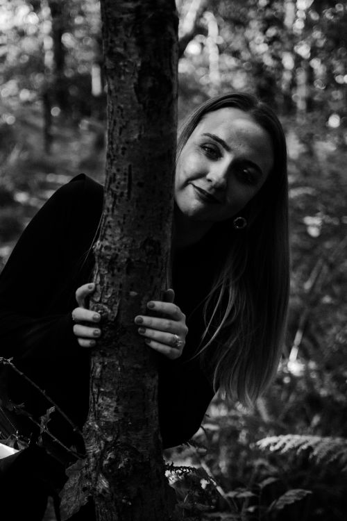 Free Black and white portrait of a woman with long hair peeking from behind a tree in a forest. Stock Photo