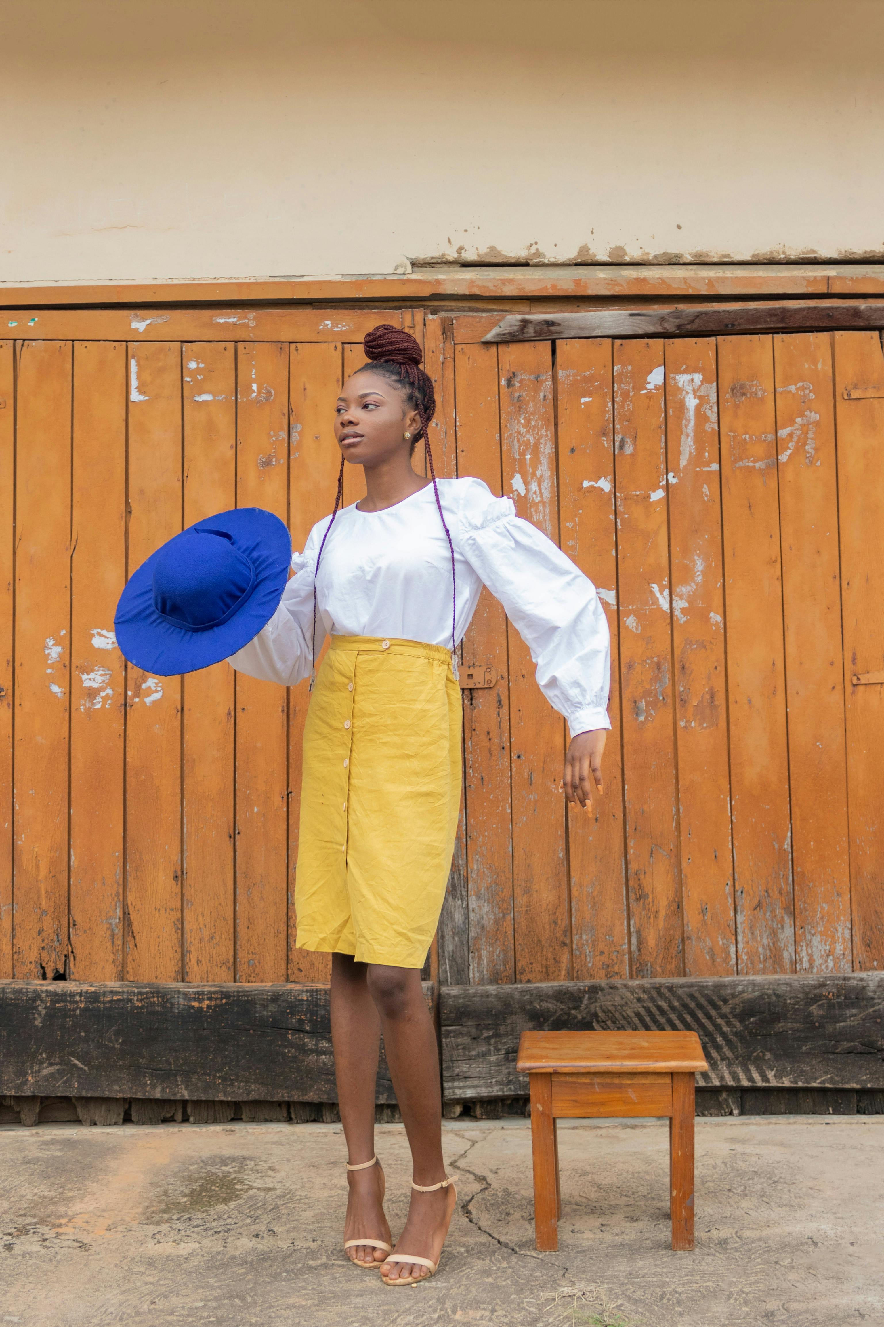 Stylish African woman in a yellow skirt holding a blue hat against a rustic wooden backdrop.