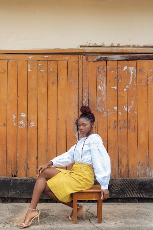 Side view of self esteem slim ethnic female with stylish hairstyle in trendy yellow skirt and high heels sitting on wooden stool near shabby wall and looking at camera
