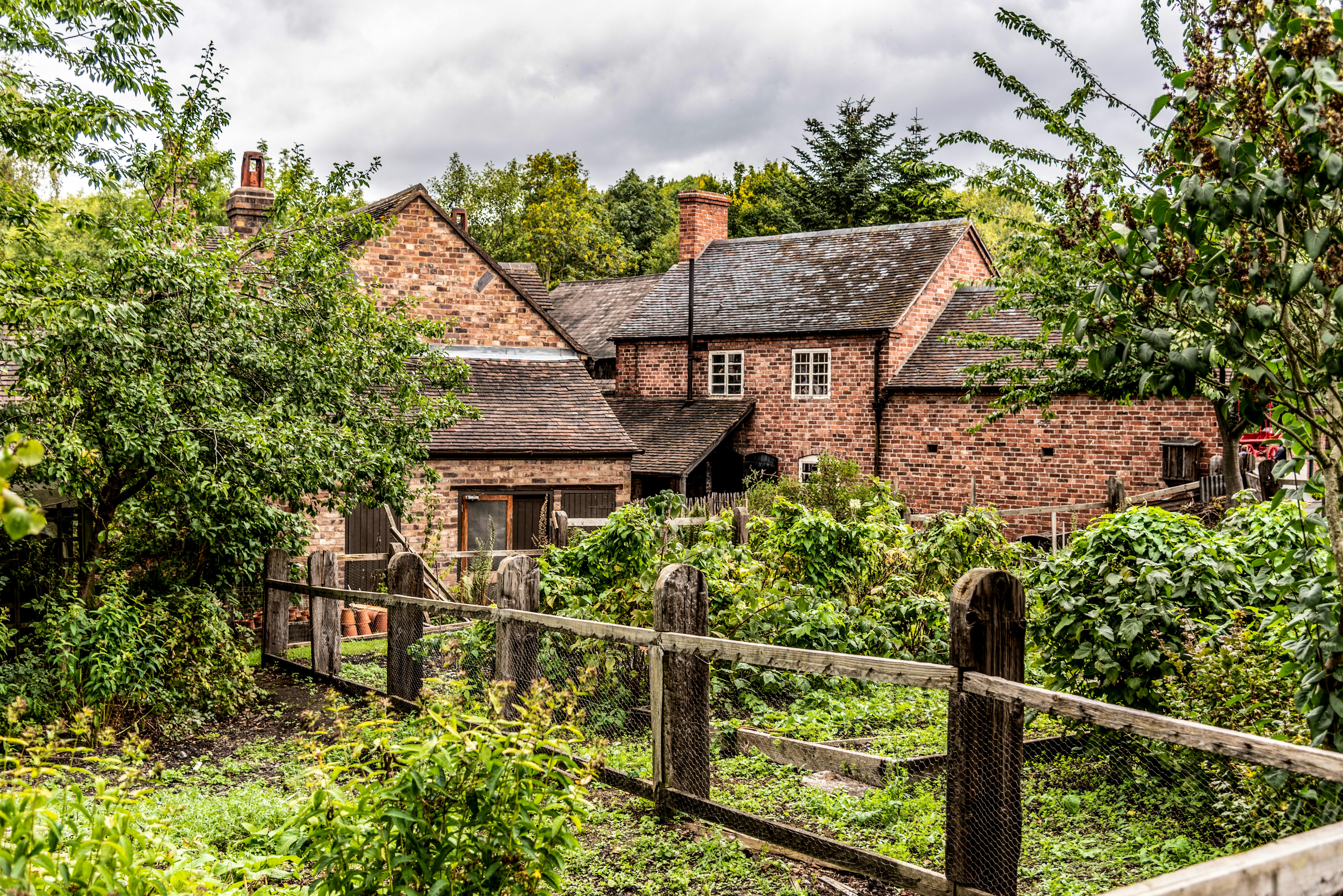 Quaint English brick cottage surrounded by a lush green garden with wooden fencing.