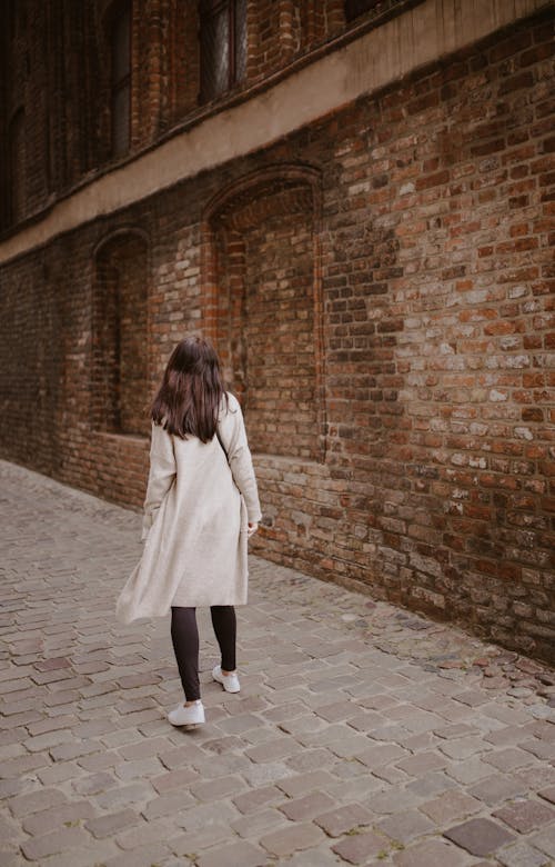 Back view full length brunette tourist in casual wear strolling along paved walkway near medieval masonry buildings in old city district