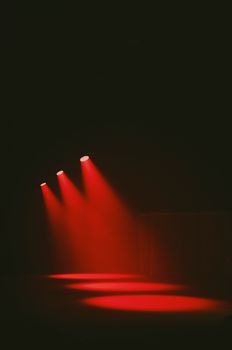 Three red spotlights illuminate a dark empty stage, creating a dramatic and moody atmosphere.