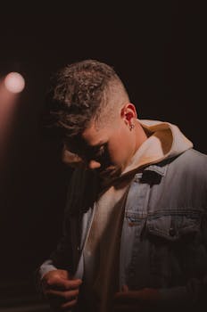 Young bored male with piercing wearing casual clothes standing in dark room and looking down