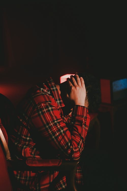 Side view of tired young guy in checkered shirt with hands behind head sitting in chair in dark room