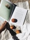 From above of crop unrecognizable traveler reading interesting book while relaxing on beach with ripe peaches on sunny day