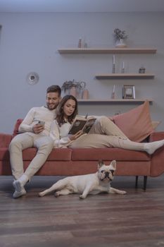 一对夫妇和一只法国斗牛犬在沙发上悠闲地待着，在室内看看书、玩玩手机