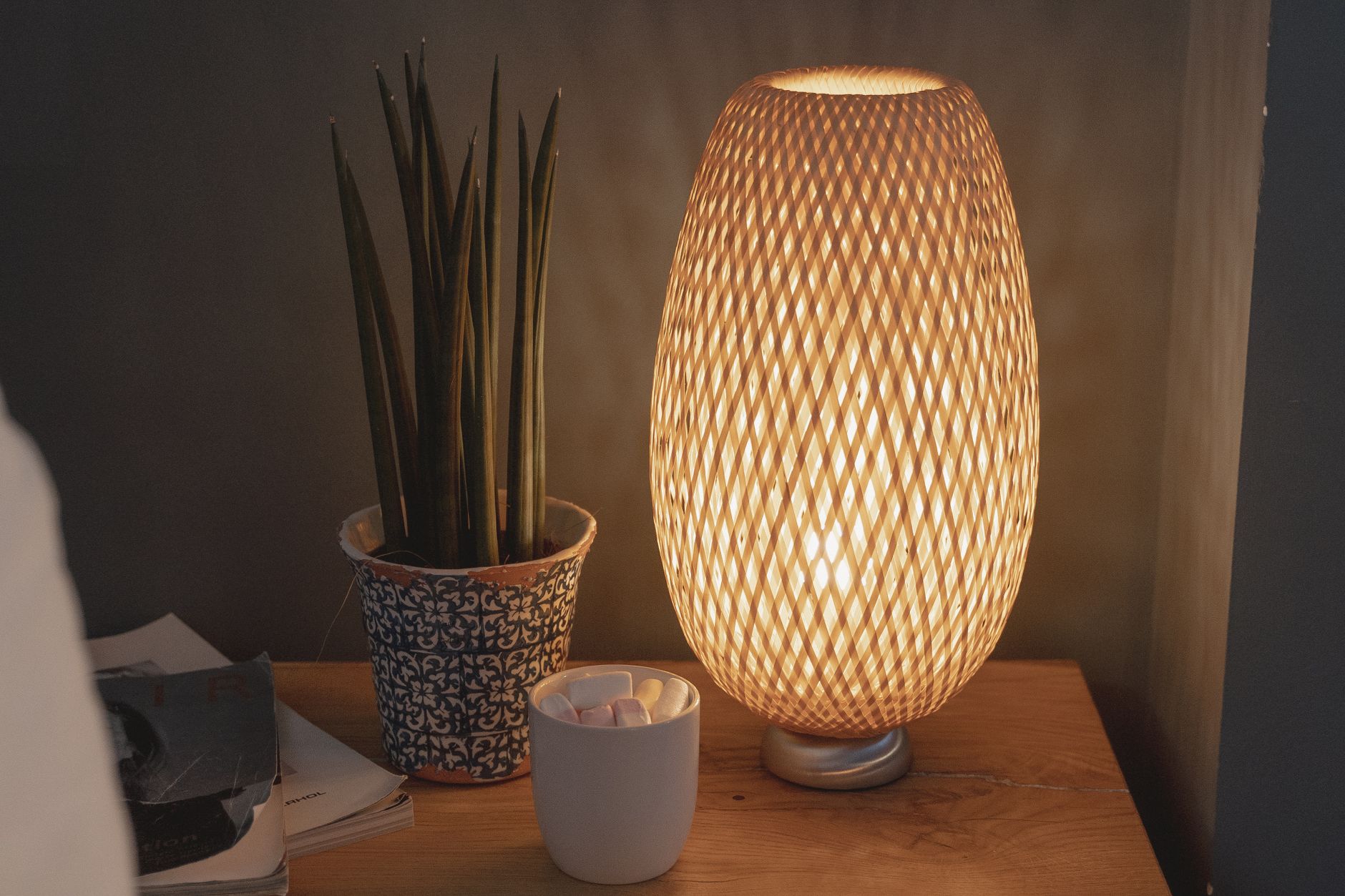 A warm, beautifully lit nightstand featuring a woven lamp and potted plant, creating a cozy atmosphere.