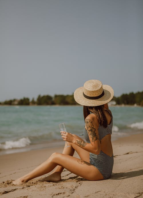 Free Unrecognizable female with tattoos and brown hair in swimwear and hat sitting on sandy beach near waving sea in sunny day Stock Photo