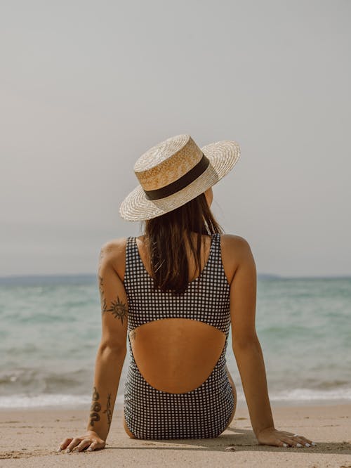 Woman with tattoos in swimsuit and hat resting on sandy beach