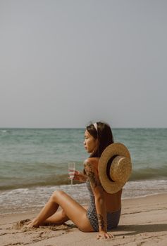 Side view of young tanned tattooed Asian female traveler in swimwear and straw hat drinking wine while relaxing on sandy beach and admiring sea