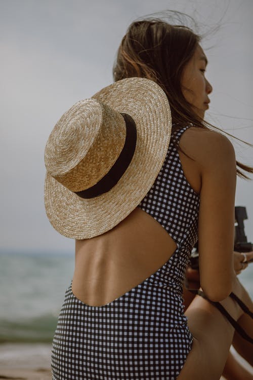 Back view of calm young ethnic female in swimsuit and straw hat relaxing on sandy beach with photo camera in hands and looking away