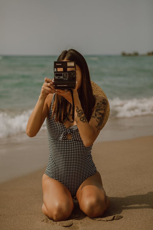 Free Unrecognizable slender lady taking instant photos on vintage camera while resting on sandy sea coast Stock Photo