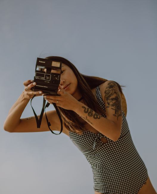 Low angle of positive young Asian female photographer with tattoo on arm in trendy swimsuit photographing on retro film camera against cloudless sky on sunny day
