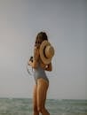 Sid view of anonymous stylish slender female traveler in swimsuit and straw hat standing on beach near waving sea during summer holidays on sunny day