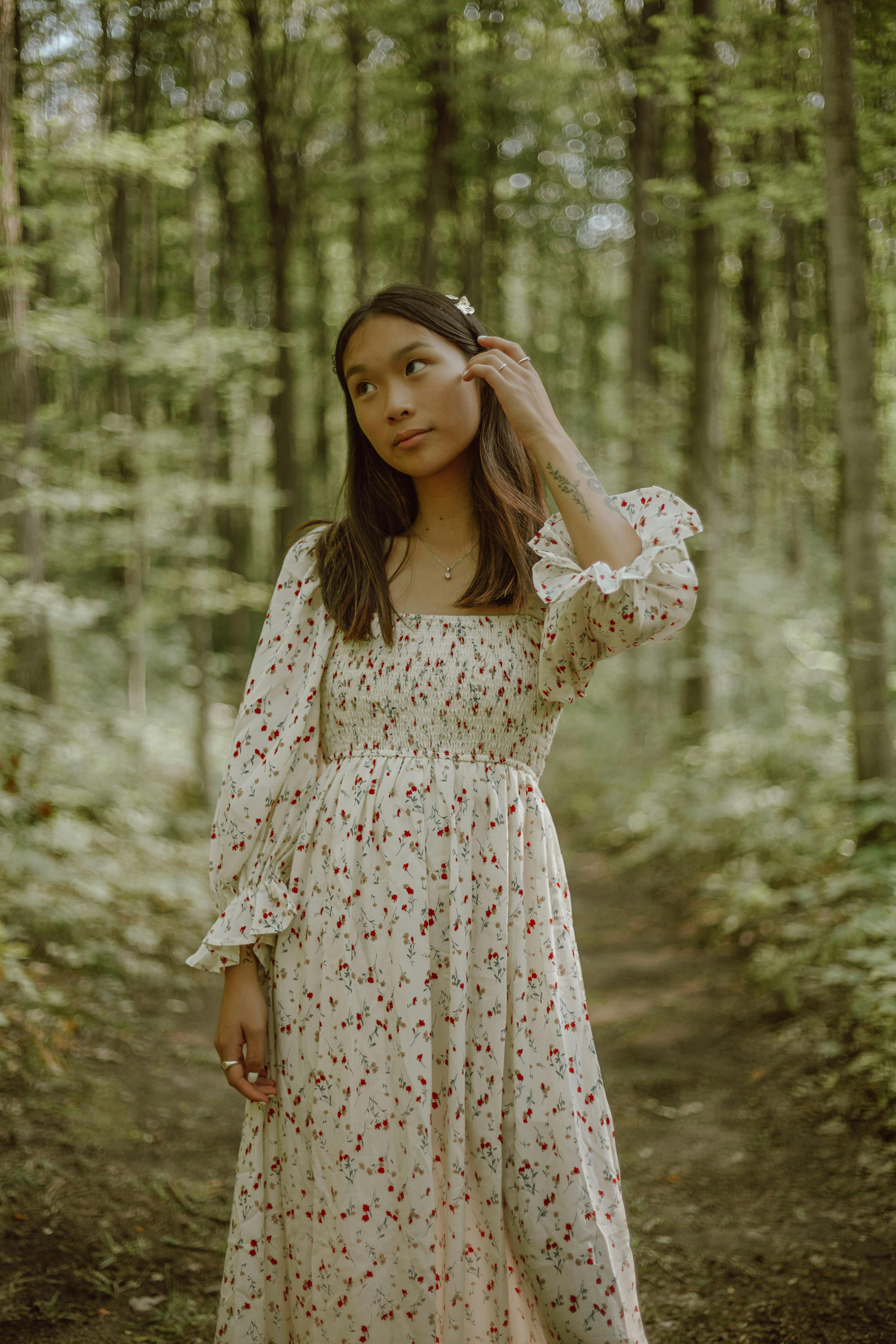Dreamy young ethnic lady in feminine dress touching long dark hair and looking away while relaxing in green forest on sunny day