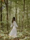 Side view of unrecognizable young slim female in romantic maxi dress walking in green forest amidst lush green trees on sunny day