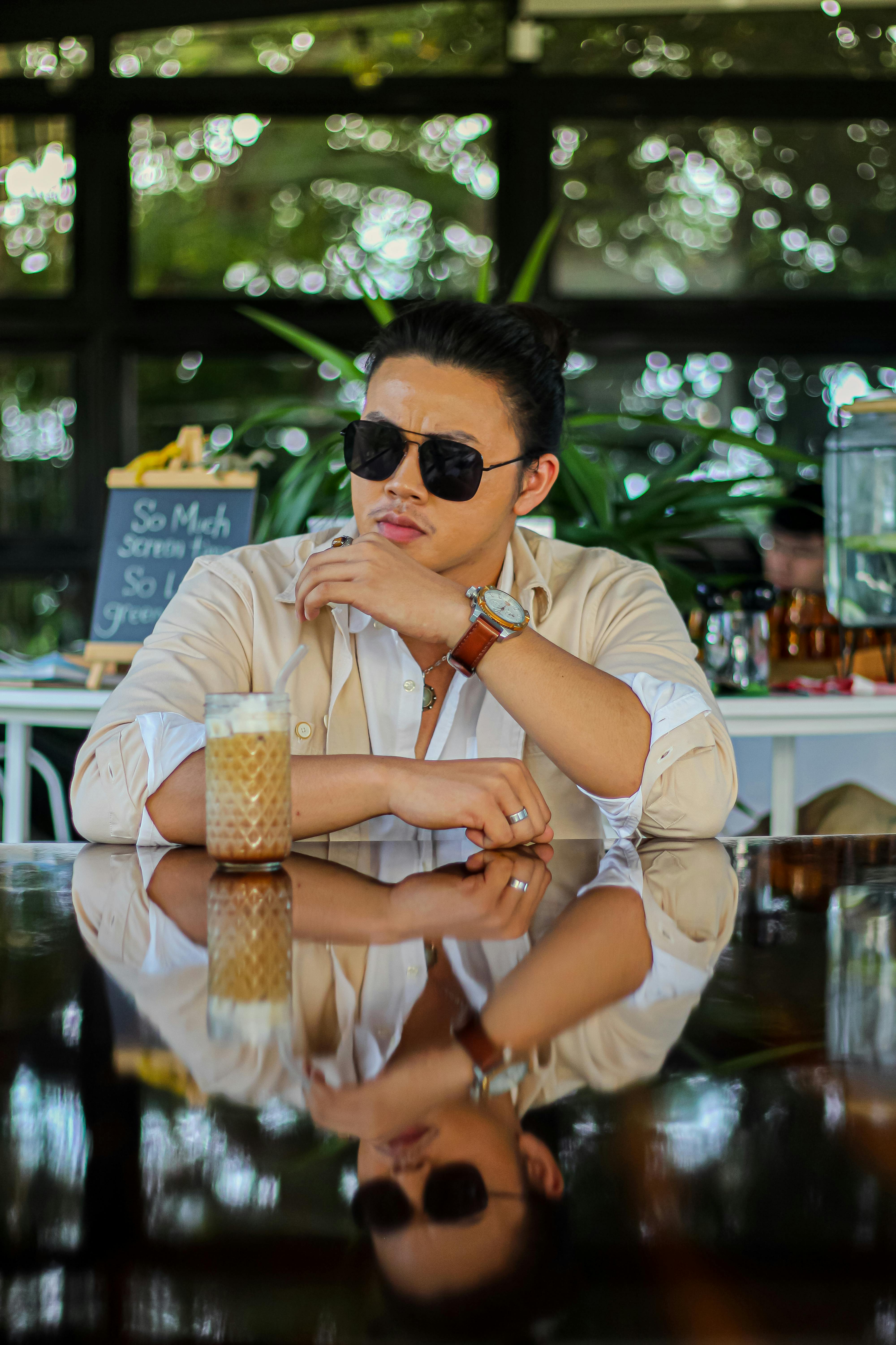A fashionable man with sunglasses enjoys a drink in a cozy cafe, reflecting on a polished table.