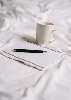 A white mug beside a spiral notebook and pen on white bedding.
