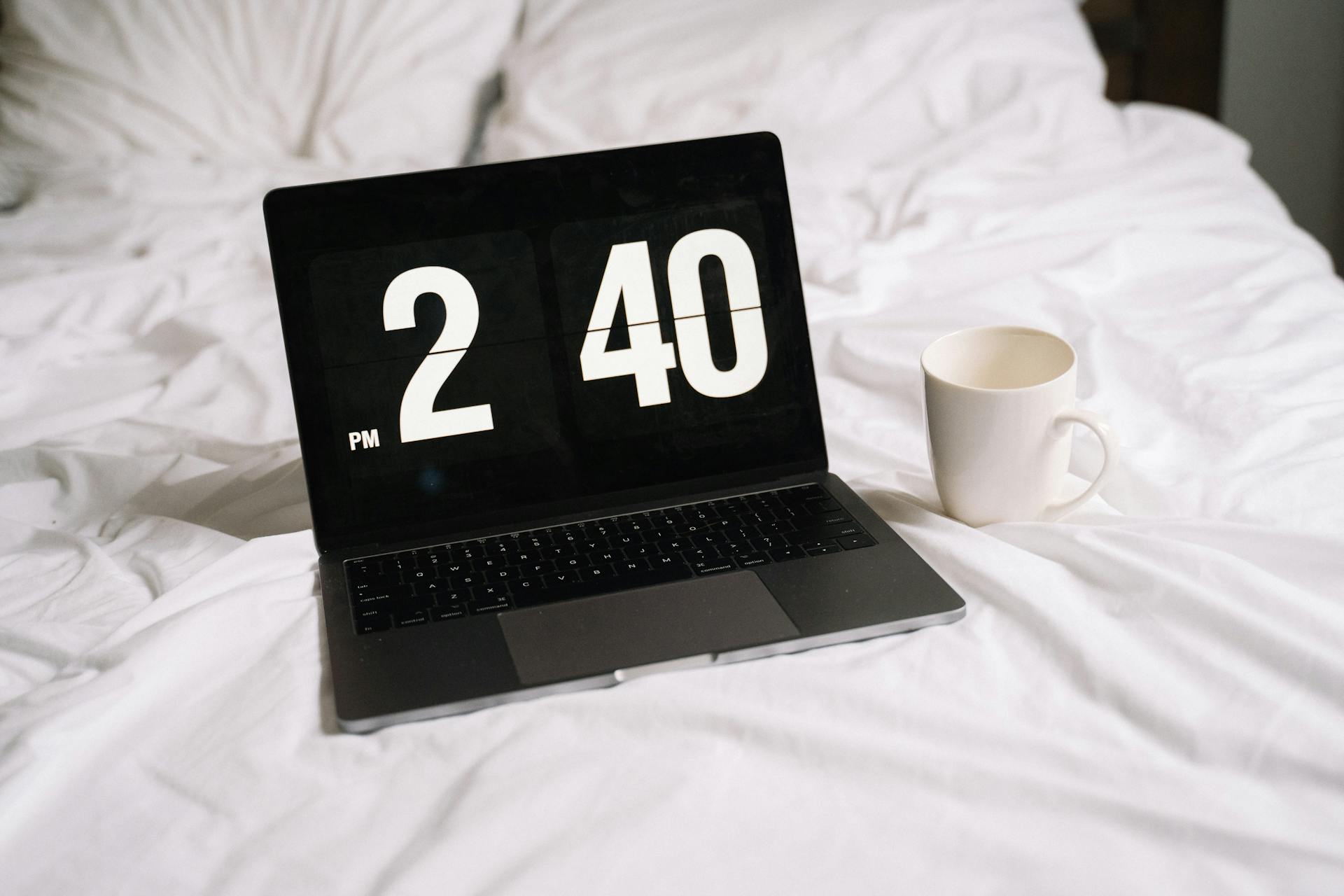 A laptop displaying time and a coffee mug on a white bed, creating a relaxed work or leisure atmosphere.