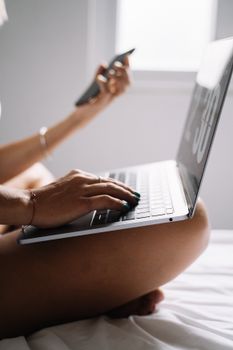 A woman multitasks with a laptop and smartphone on her lap in a brightly lit room.